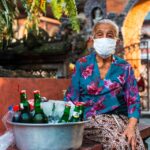 Senior Balinese street vendor in Ubud, Bali wearing a mask and traditional attire with beverages for sale.